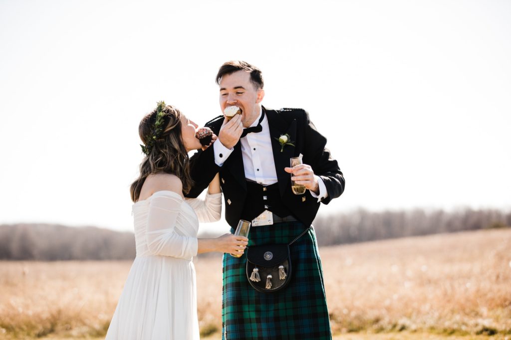 A couple eats cupcakes after their elopement. She is wearing a white wedding gown, he is in a kilt. 