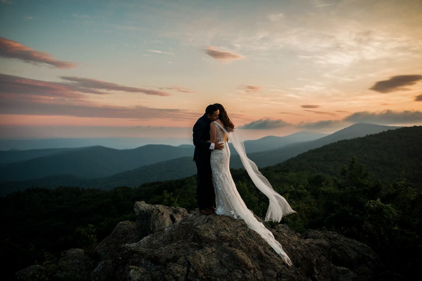 A couple stands on top of a mountain in Shenandoah National Park. The wind is blowing the bride's veil