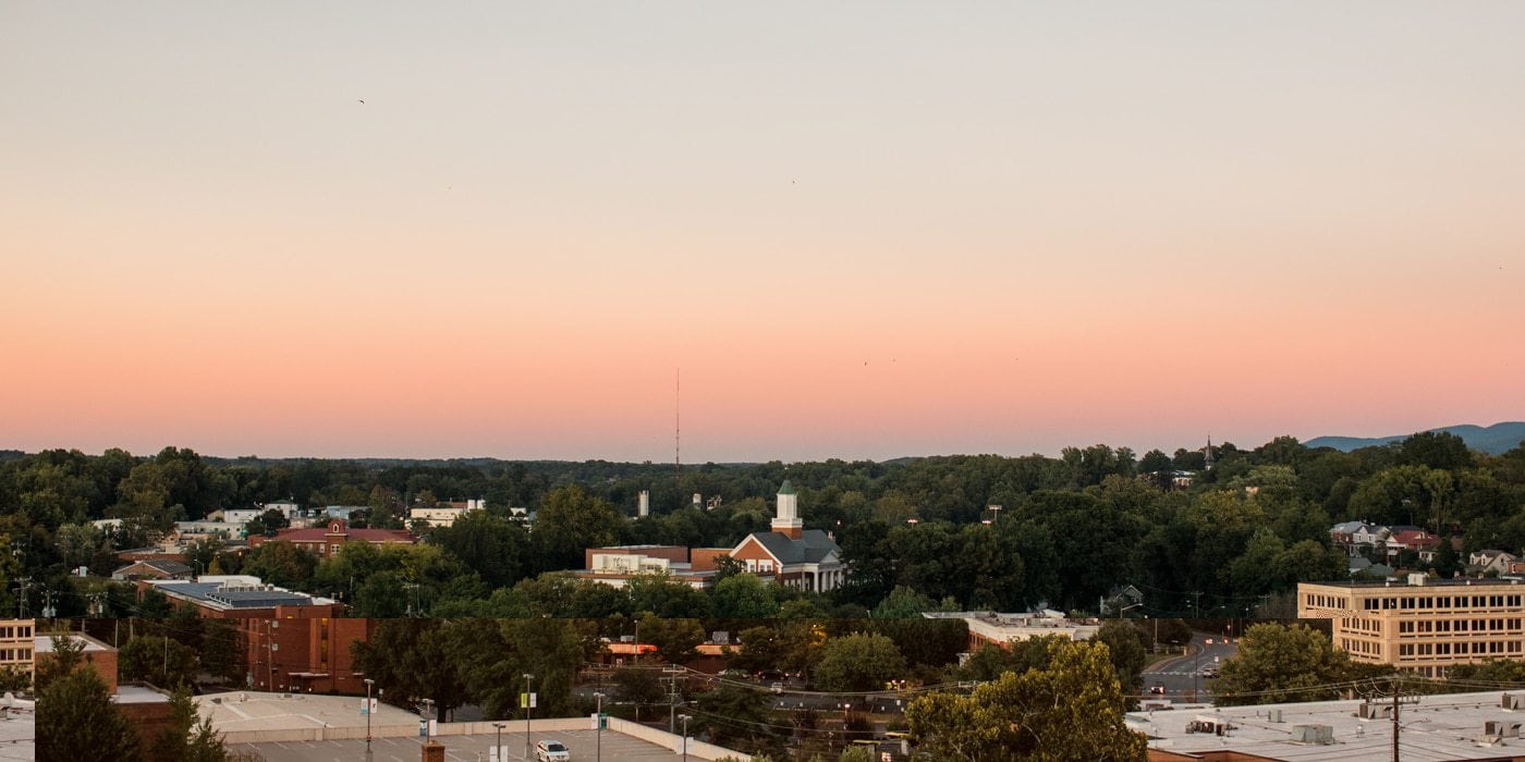 A skyline view of Charlottesville at sunset. The sky is pink and buildings can be seen in the distance