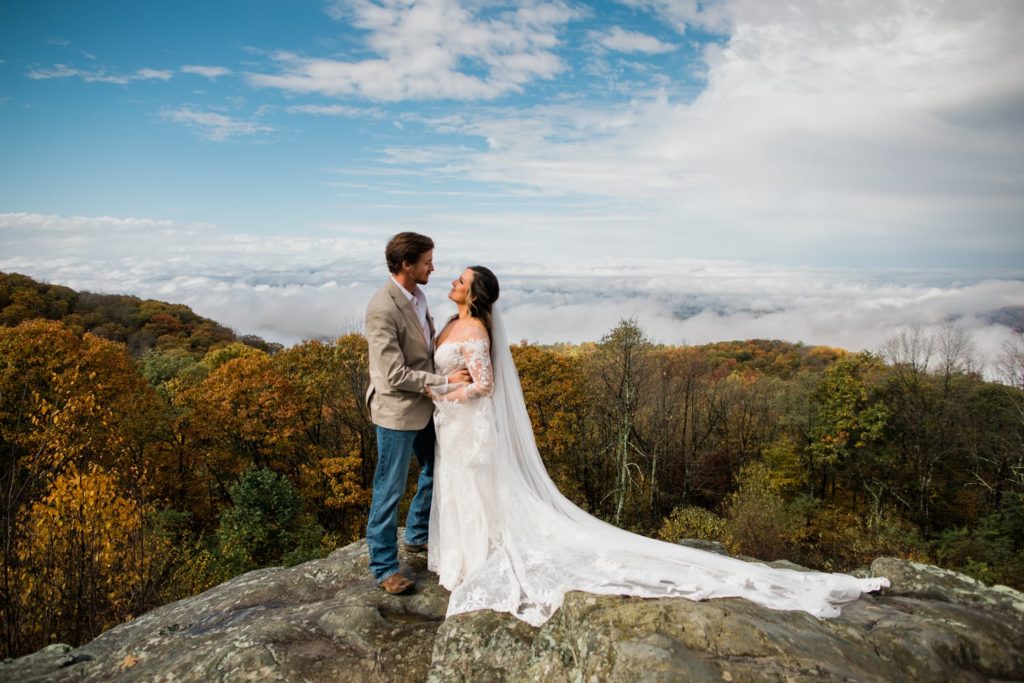 A bride and groom embrace and look at each other in Shenandoah National Park. It's fall and the leaves are orange. They are on top of a rock on Skyline Drive and cloud inversions can be seen in the backgorund 
