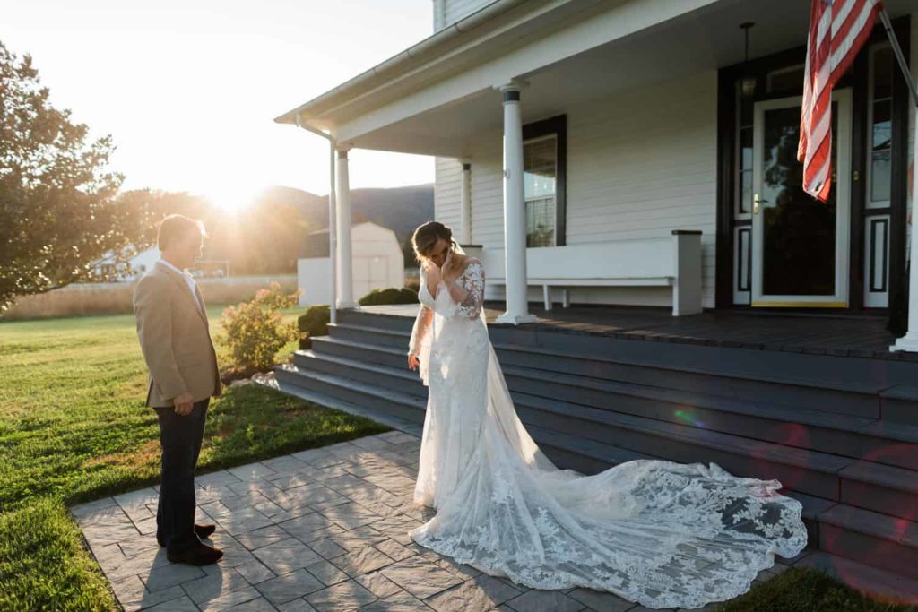 A bride wipes tears from her eyes after seeing her dad on her wedding day. She is standing in front of a farmhouse Airbnb. An American flag is dancing in the wind and the sun is about to set behind the mountains 