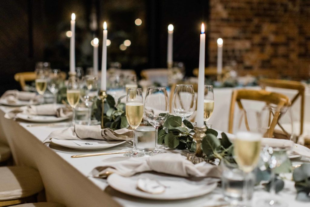 A table scape from an intimate wedding at The Wool Factory. Candles are lit, eucalyptus is being used as. a runner and wine glasses are filled with white wine. 