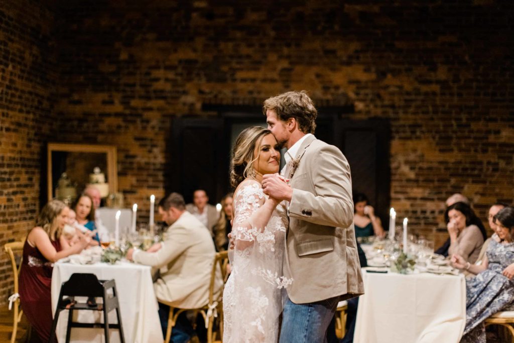 A bride and groom slow dance during their intimate reception at the wool factory while their guests watch from seated tables. 
