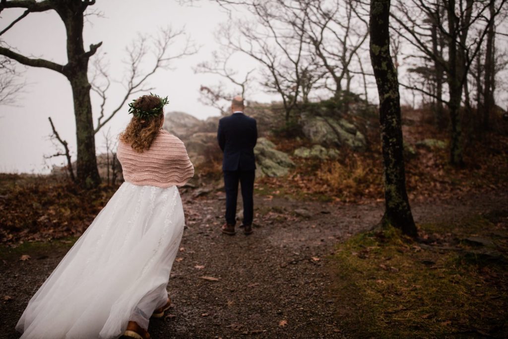 A bride in a white dress and flower crown walks toward her groom for a "first look" on top of the mountain 