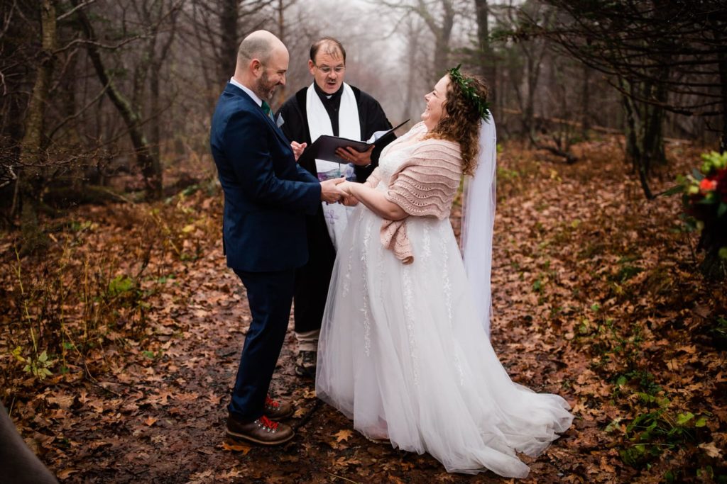 A bride and groom laugh during an elopement ceremony in Shenandoah National park in the fall. There are leaves on the ground and fog hanging in the trees