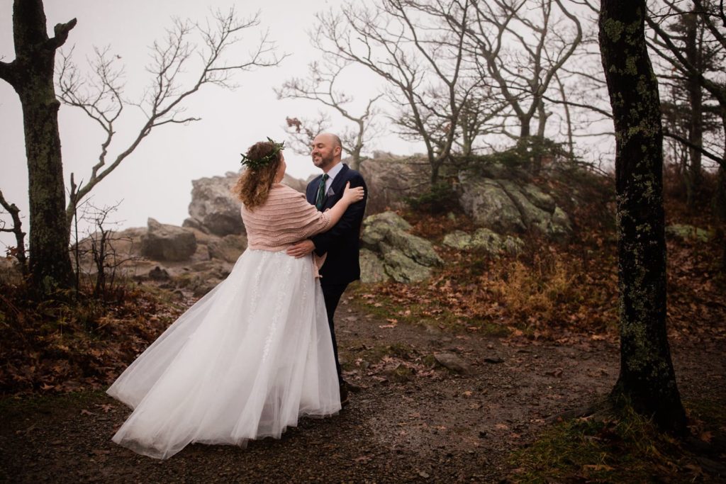 A bride and groom smile excitedly at each other after a "first look." They are on top of a mountain that is covered with fog