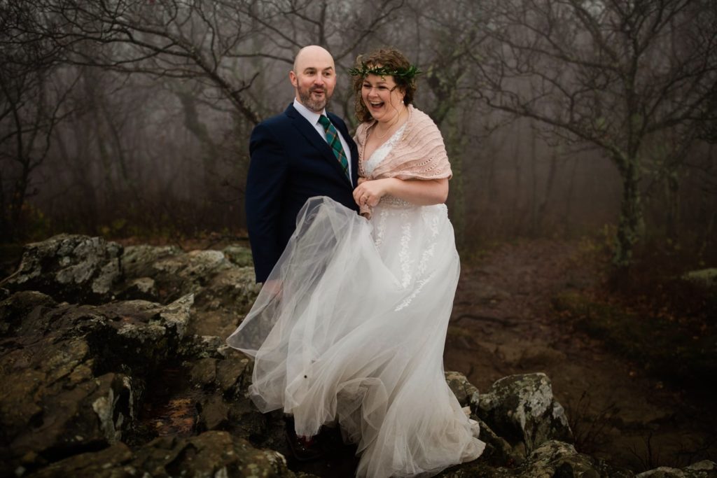 bride in a flowy dress stand beside her groom who is making a goofy face. They are standing on top of a mountain in Shenandoah National Park and fog is around them