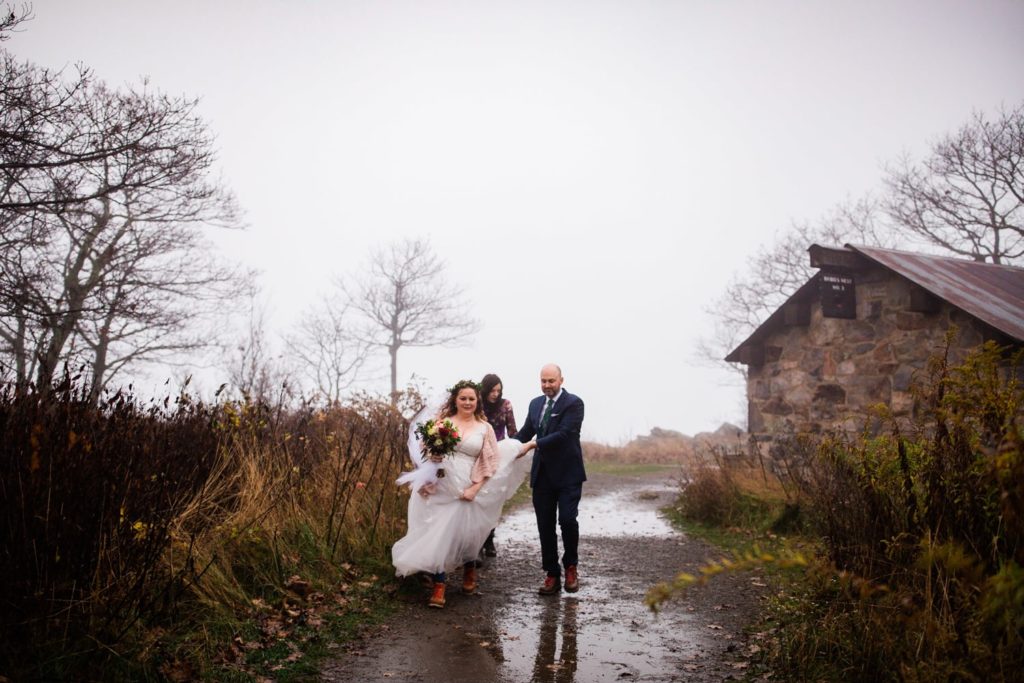 A bride, groom and friend walk to the ceremony on top of a mountain. THe groom is holding the bride's dress as they walk around a puddle 