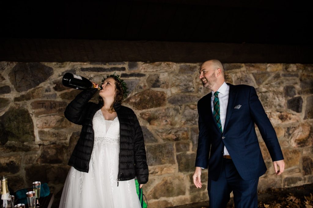 A bride chugs champagne from the bottle while her groom looks on and laughs during an elopement celebration