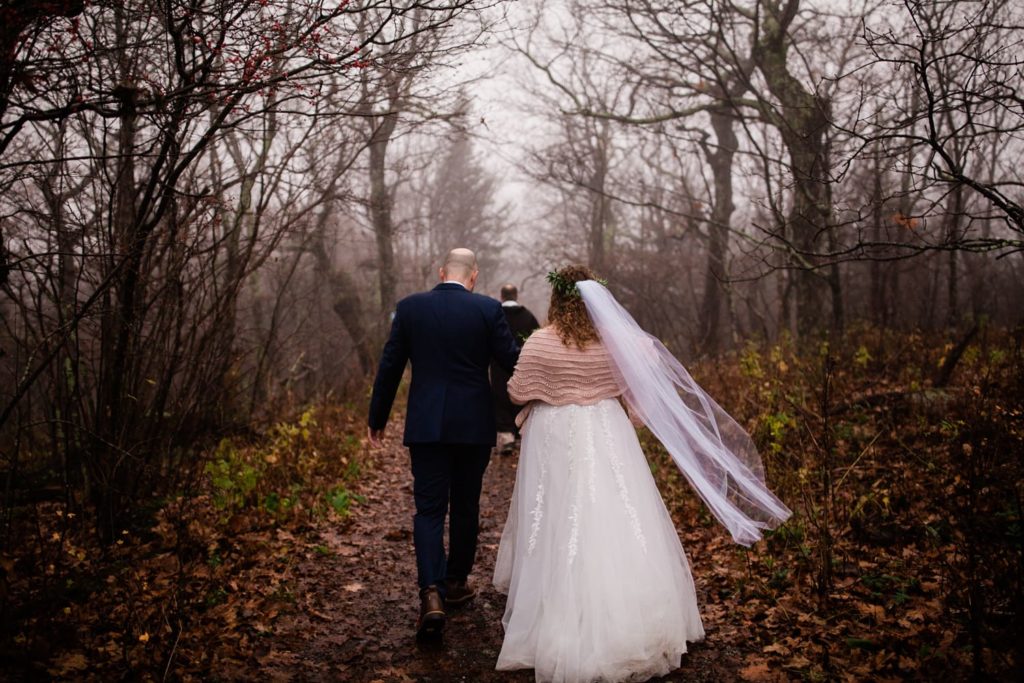A bride and groom walk away from the camera down a mountain trail -- she is wearing a pink shawl and her veil is blowing in the wind 