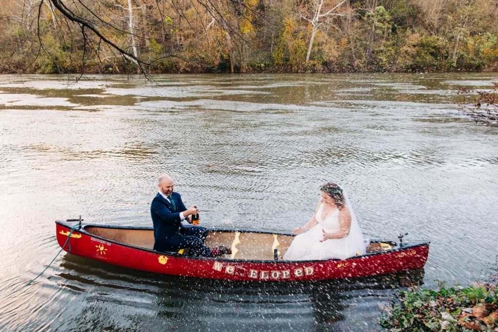 A bride and groom sit in a red canoe on the Shenandoah River that is decorated with a "we eloped" sign. The groom is popping champagne and accidentally sprayed his bride 