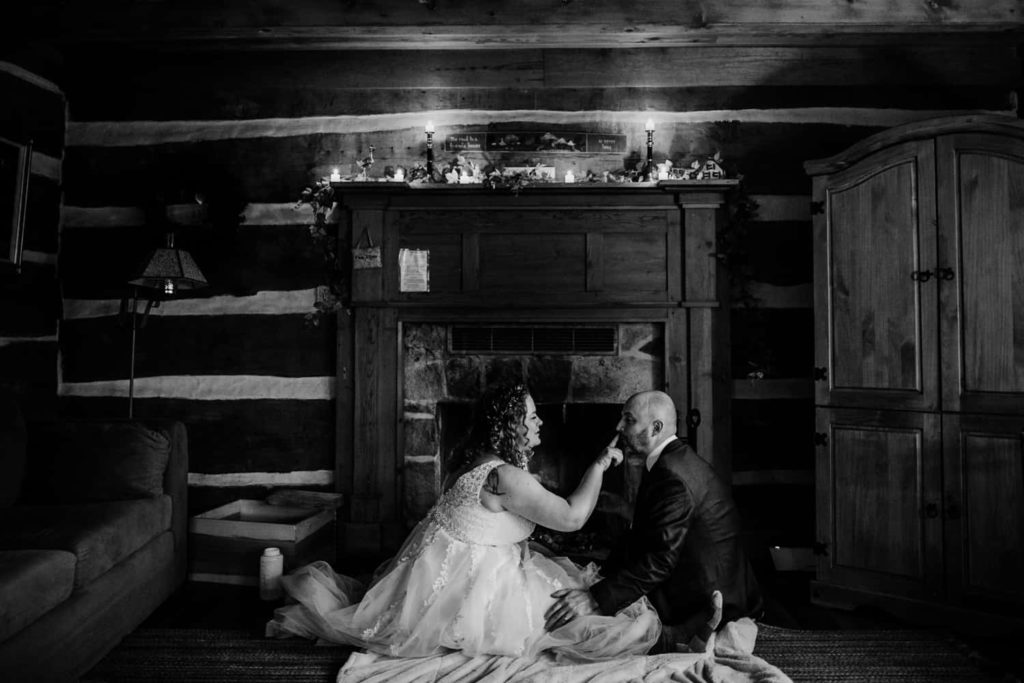 A bride and groom feed each other s'mores beside an indoor fireplace 
