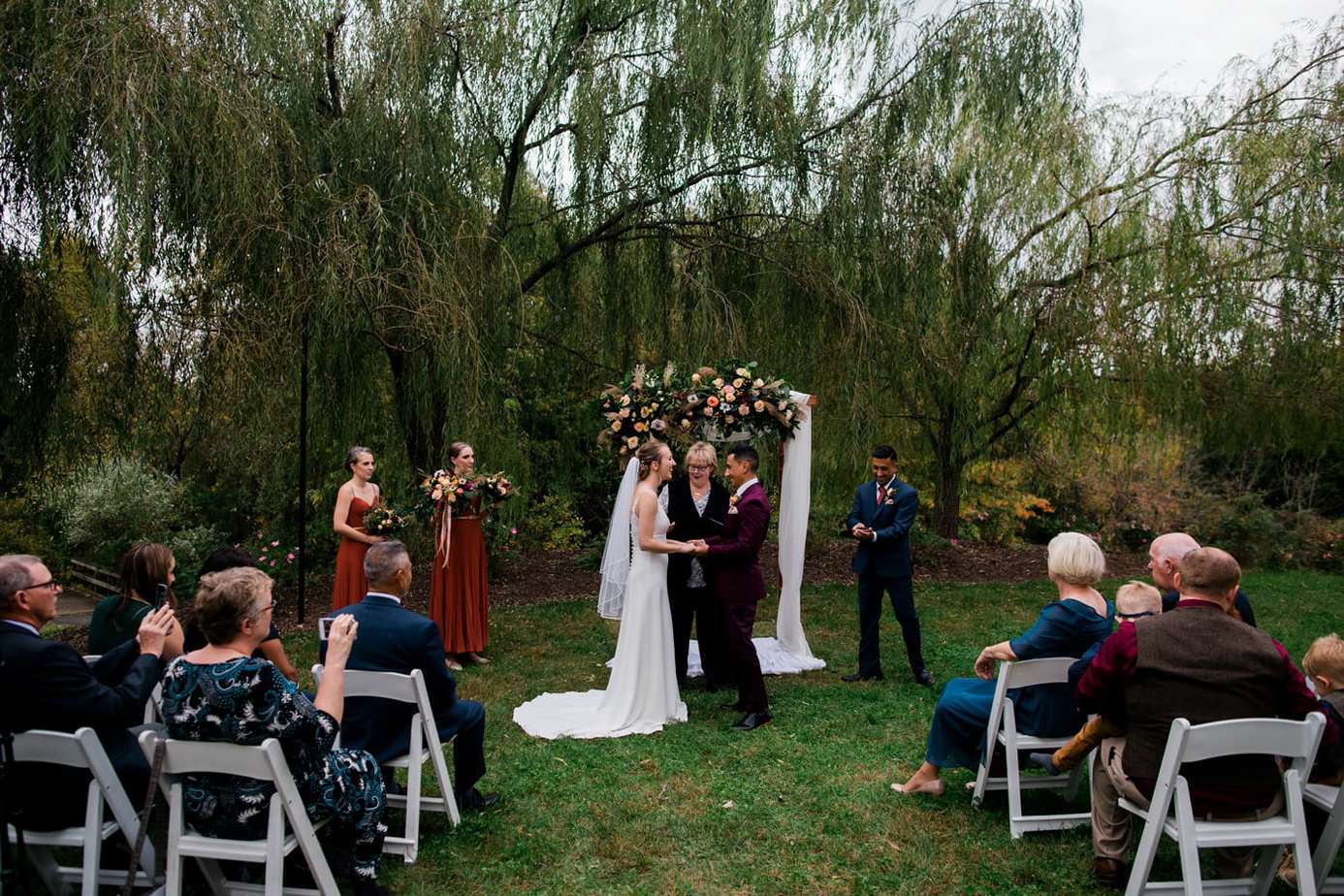 A bride and groom laugh during their wedding ceremony at Clyde Willow Creek Farm. Willow Trees are behind an arbor decorated with flowers and white cloth. Guests are seated in white chairs and there are two bridesmaids and a groomsman.