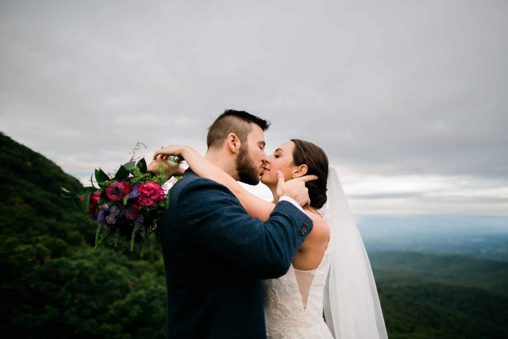 A bride and groom kiss in front of blue green mountains. The bride's arms are wrapped around the groom's neck and her bouquet dangles 
