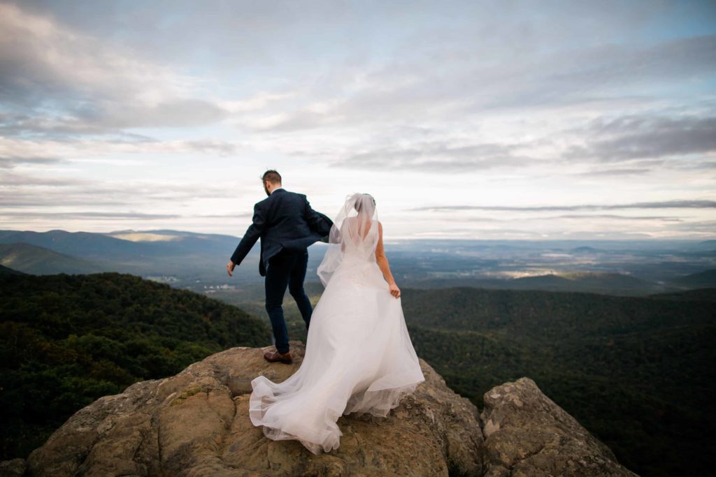 The wind blows a groom's jacket and bride's dress on a rocky mountain scene. 