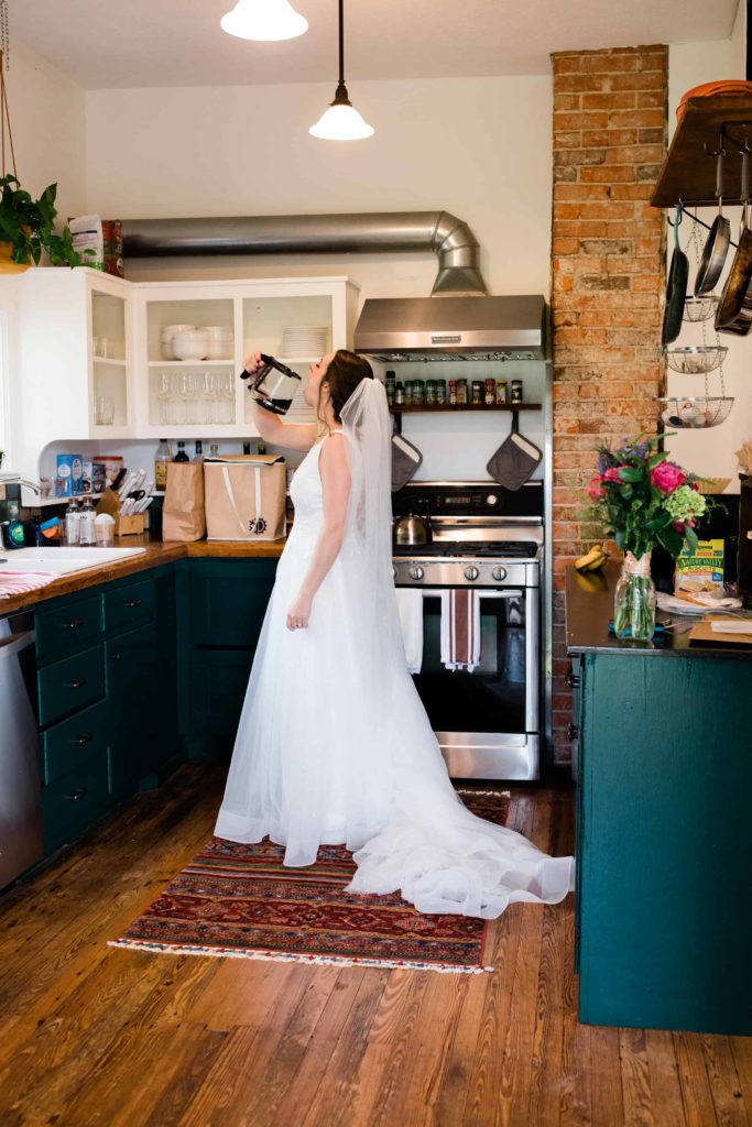 A woman wearing a wedding dress is standing in a kitchen. She pretends to pour coffee into her mouth directly from the coffeepot. 