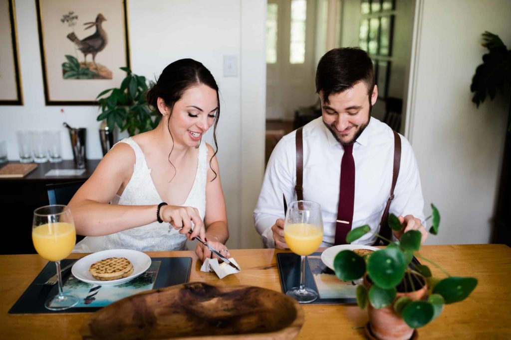 A woman wearing a wedding dress and a man wearing a tie and suspenders sit at a table together. They have mimosas and frozen waffles on the plate 