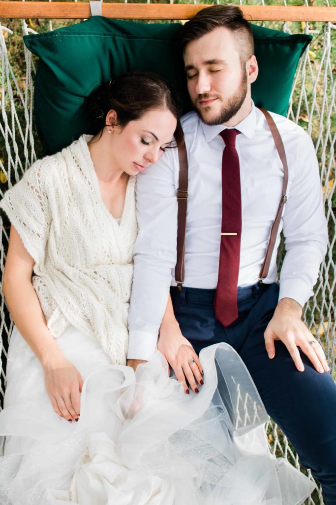 A man in a suit and a woman in a wedding dress lie in a hammock and cuddle together. 