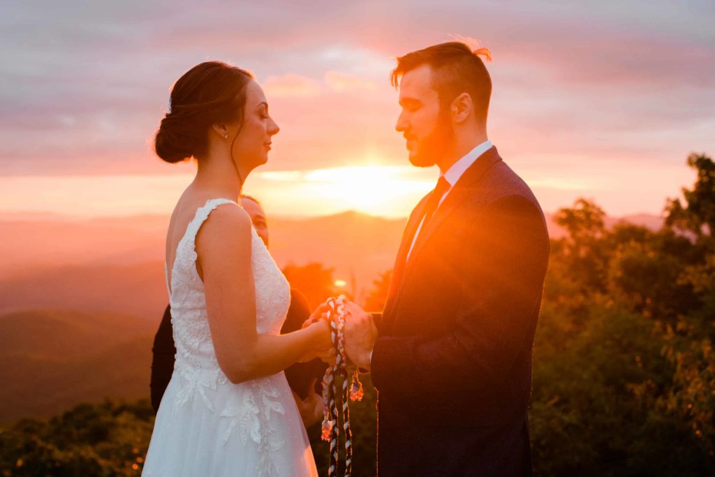 Golden orange sun shines through a couple. They are closing their eyes and holding hands. There is a rope wrapped around their hands as part of a handfasting ceremony