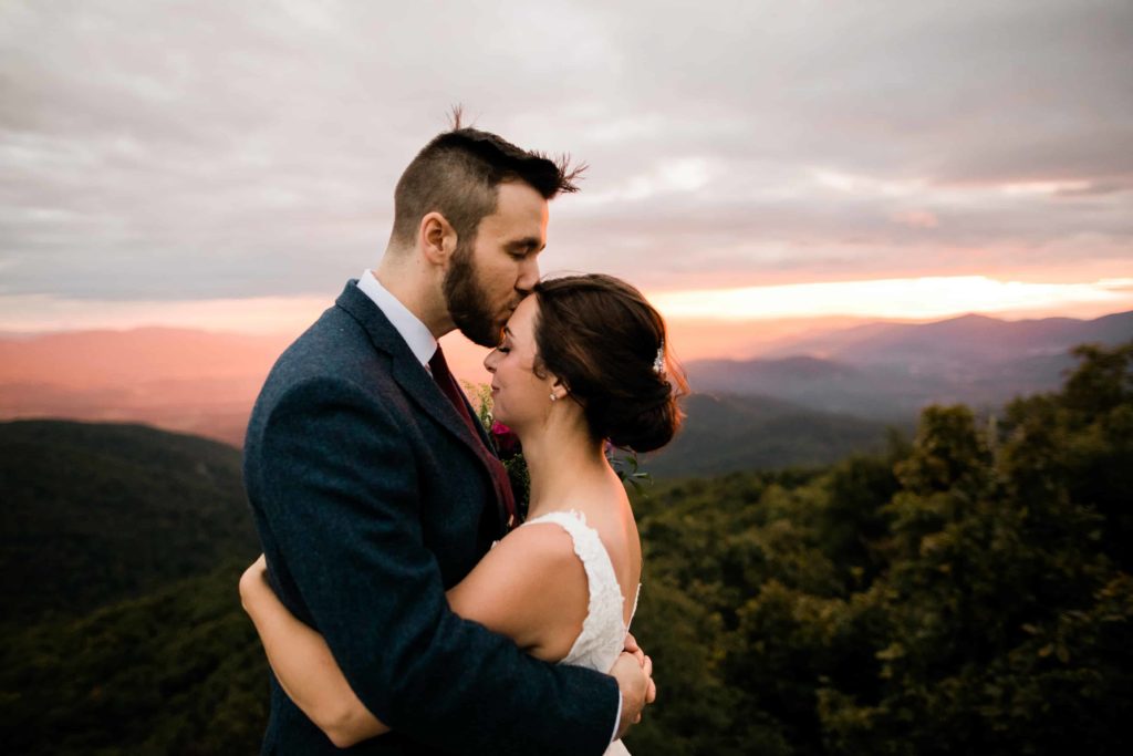 A groom kisses his bride's forehead after their elopement. The sunrise is coming up behind them 