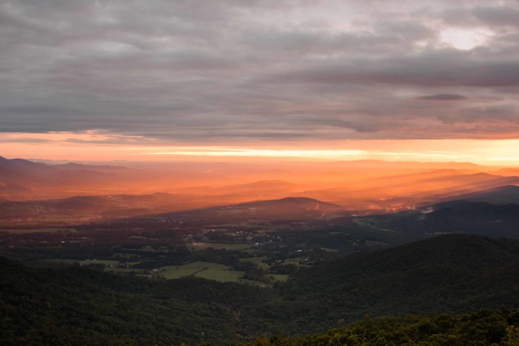 The sunrise over the Blue Ridge mountains. The top of the image is cloudy and the yellow orange sun is streaming through the bottom of the clouds over the mountains 
