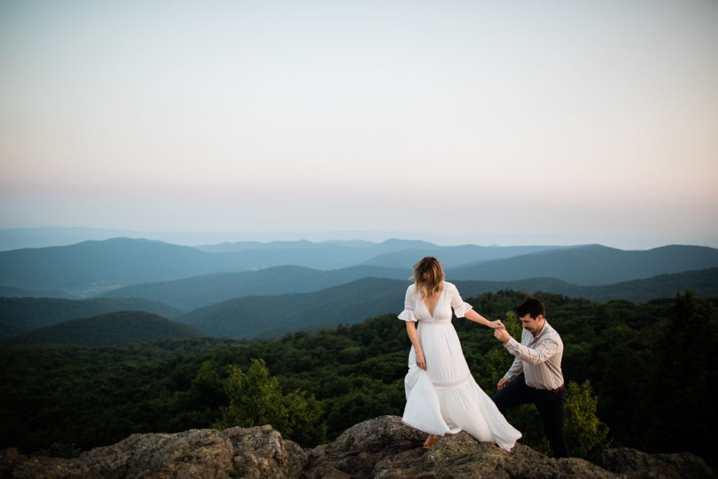 A woman in a white dress leads a man up a rocky summit. Blue Mountains are in the background