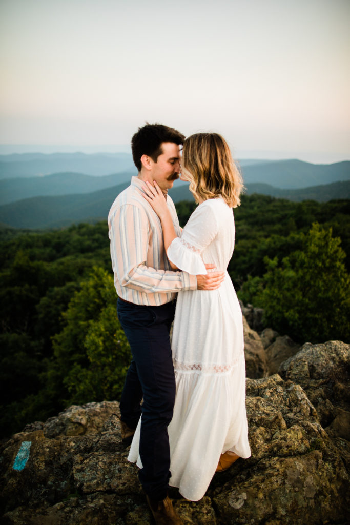 A woman in a white dress kisses a man in a striped shirt's nose. They are standing on a rocky summit and blue mountains are in the background