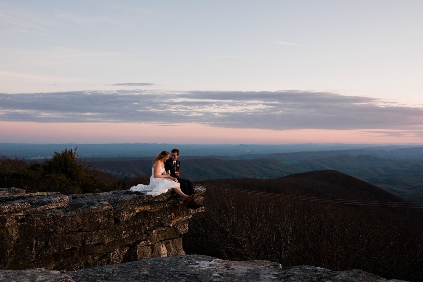 A woman and man wearing wedding garb sit on a cliff drinking champagne. A sunset is in the sky behind them and mountains can be seen in the distance