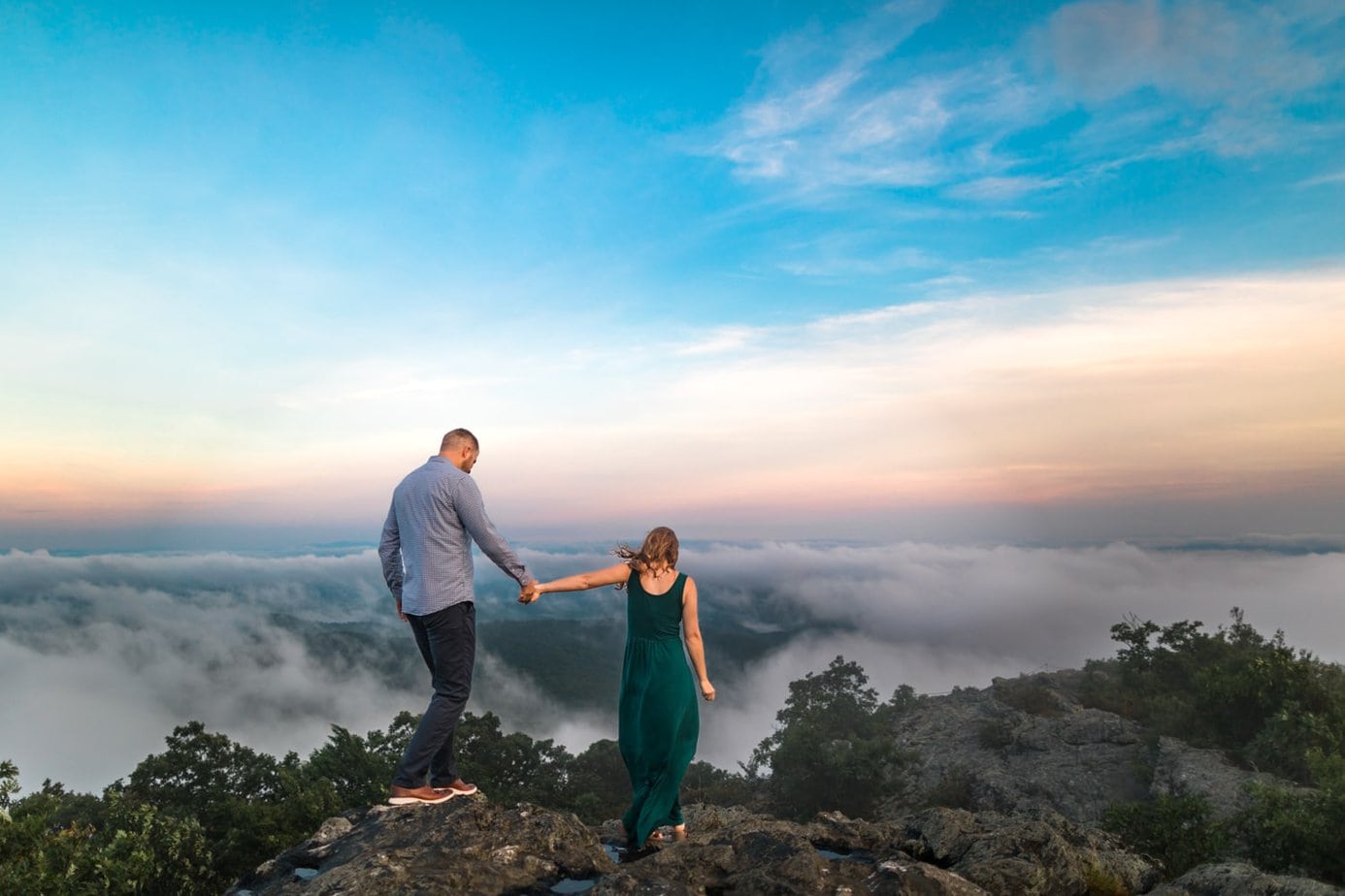 A man wearing a blue shirt and a woman wearing a green maxi dress walk away from the camera toward a mountain view. The sky is blue and fog is hanging in the mountains