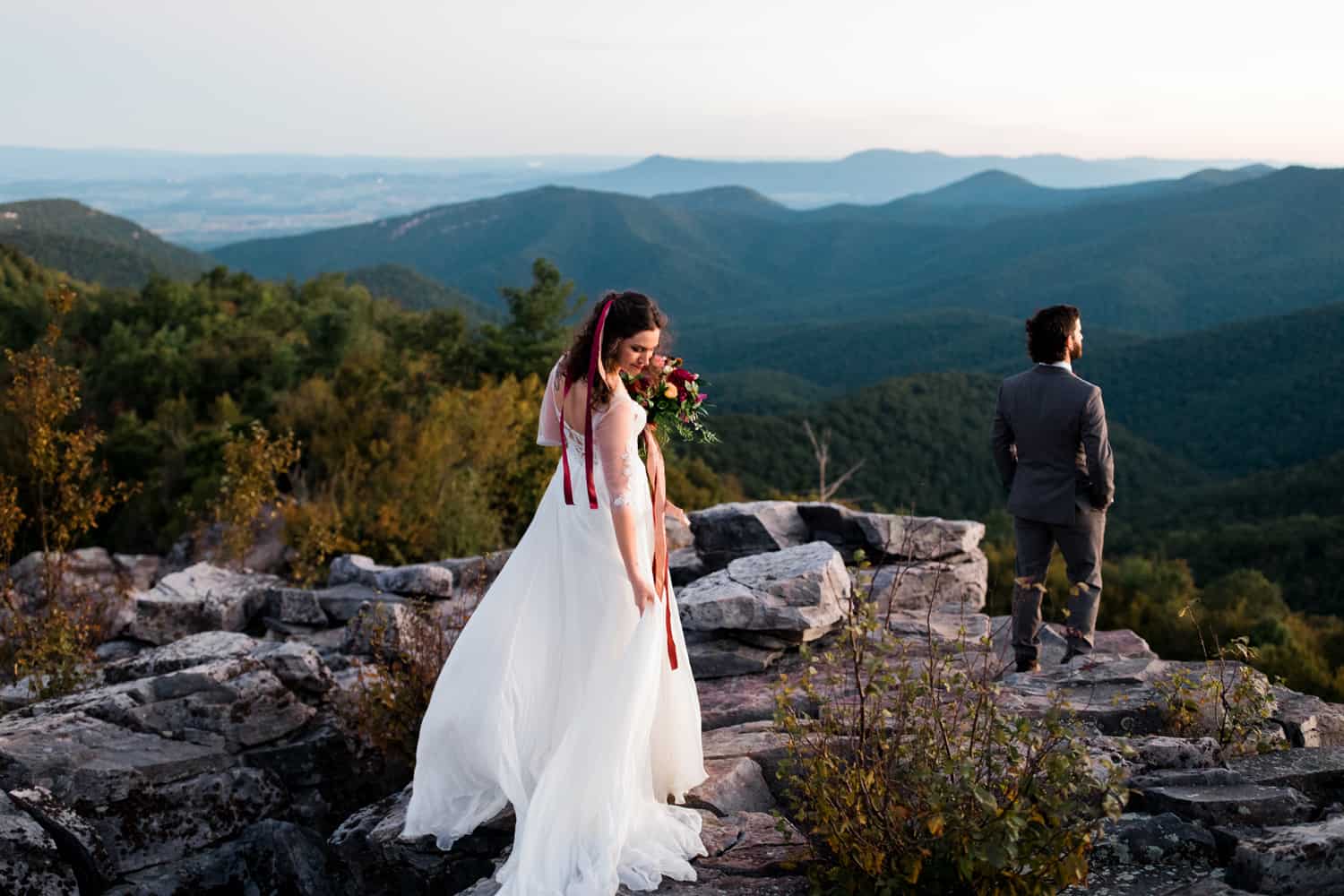 A woman in a white wedding dress looks over her right shoulder as she picks up her dress. Her groom waits for her facing out toward the mountains. Blue mountains are in the background and they are in a national park.