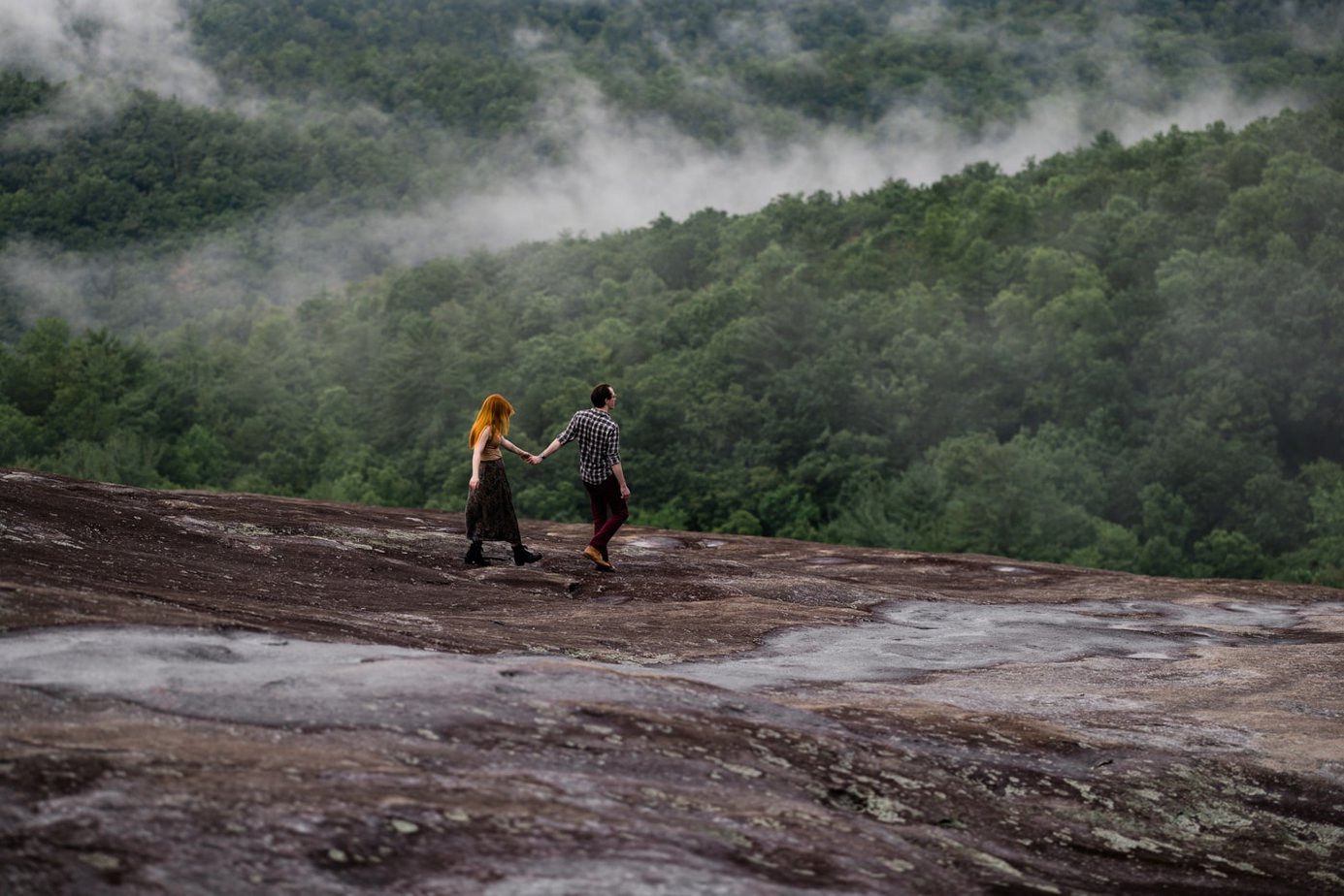 A couple walks down a rocky mountain top holding hands. Behind them fog lingers in green trees
