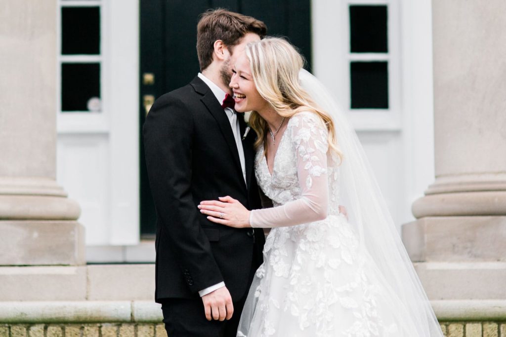 A couple laughs and embraces. She is wearing a long-sleeved white wedding gown and veil. He is in a black suit