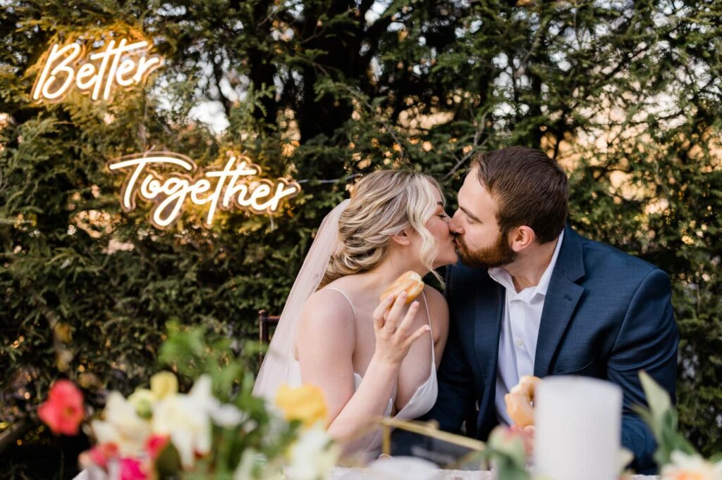A couple in wedding outfits kiss in front of a neon "better together" sign as part of  a romantic garden wedding editorial shoot in Roanoke