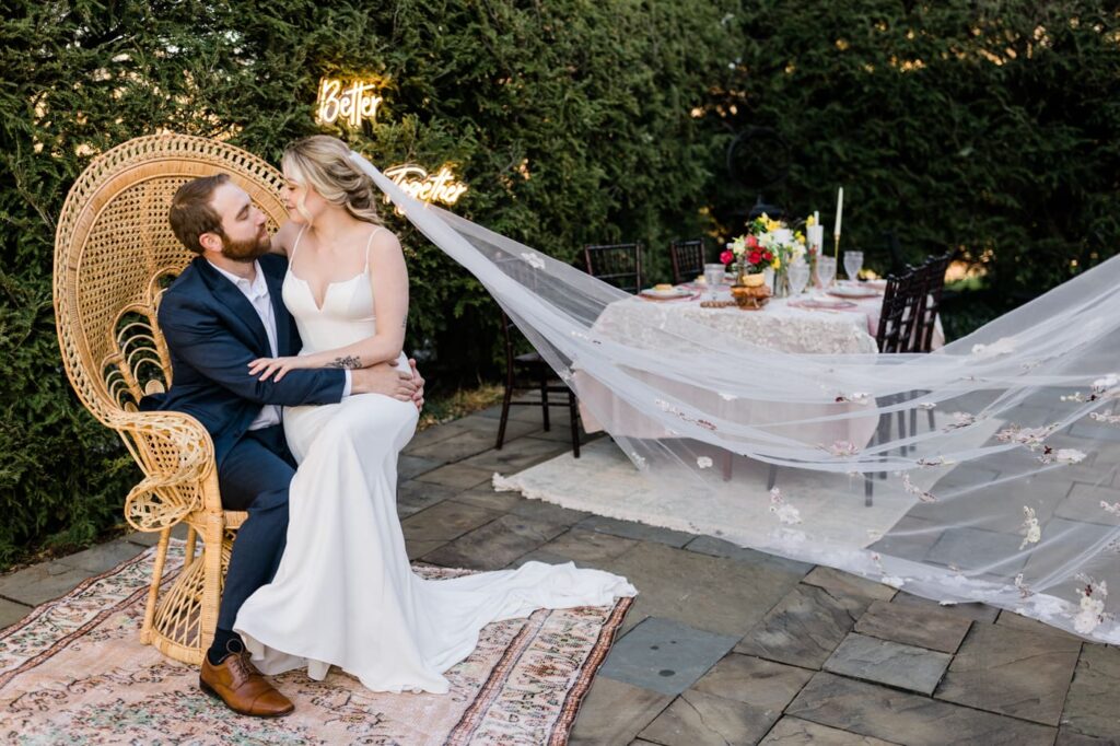 A couple sits on a peacock chair beside a tablescape. Her floral veil is flowing in wind as part of  a romantic garden wedding editorial shoot in Roanoke