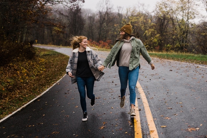 A lesbian couple runs along Skyline Drive looking at each other during their engagement session. 