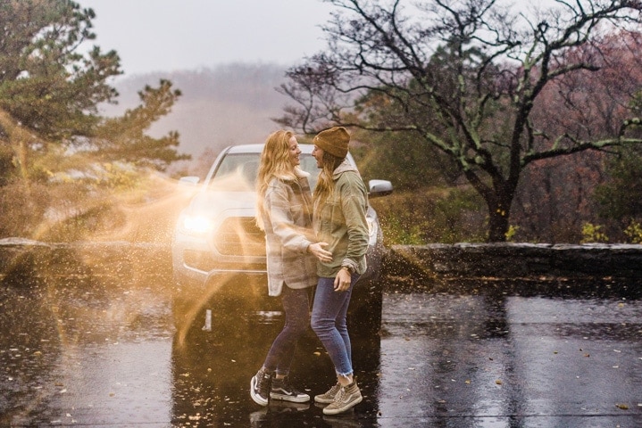 Two women embrace in front of their truck. It's raining and the headlights are on. They are doing an engagement photoshoot.