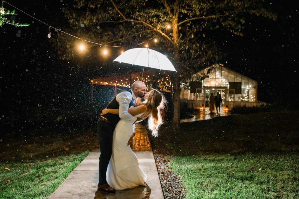 A couple dip kisses with an umbrella over their heads. It is raining and the umbrella is filled with light. 