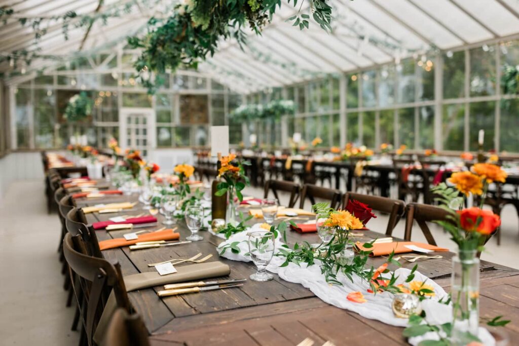 A tablescape in a greenhouse wedding venue shows lots of colorful flowers and greenery