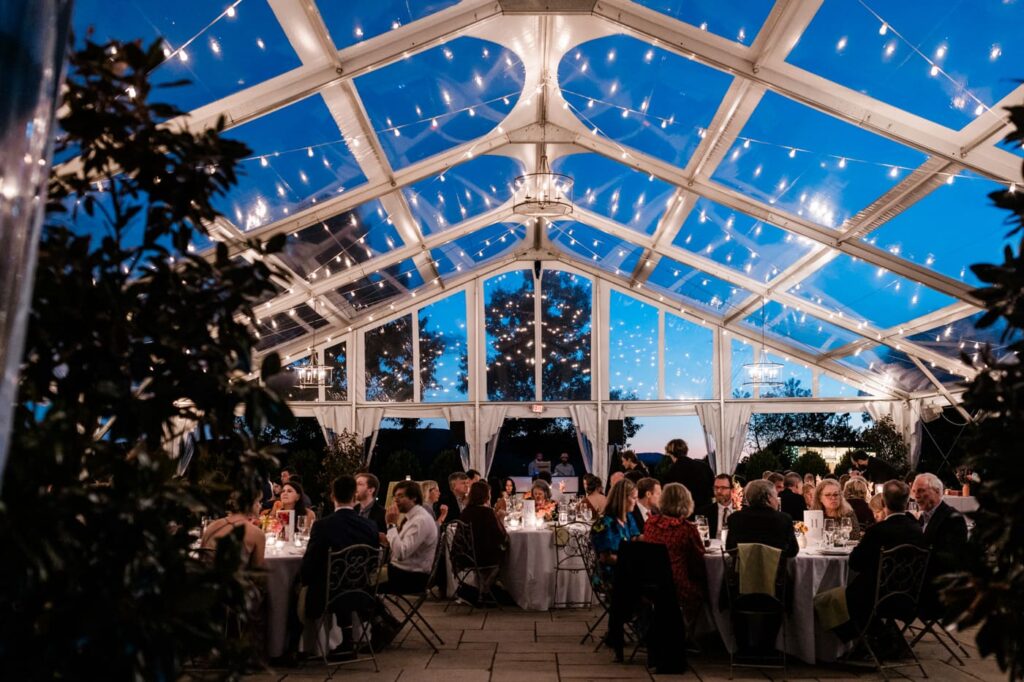The Market at Grelen, a greenhouse wedding venue, is shown at night with twilight visible through the clear tent