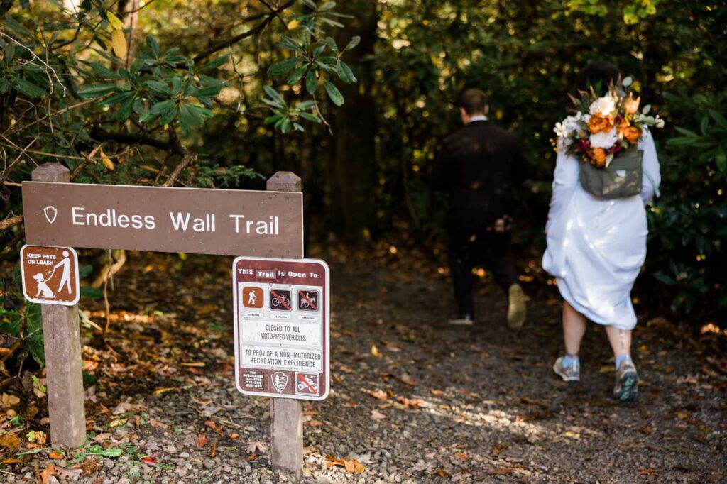 A couple wearing wedding attire walks past the sign for Endless Wall Trail at the New River Gorge